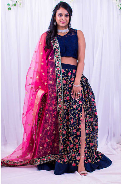 Two piece lehenga with high slit in blue skirt, covered in pink flower embroidery and gold stems. Paired with lacy blue blouse. Finish this look by draping hot pink dupatta with flower border on shoulders/ shoulder.
