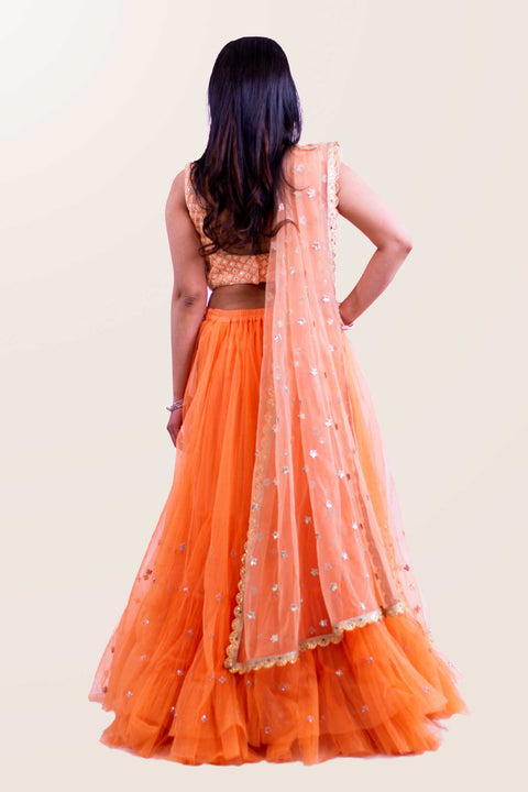 Exquisite two piece peach lehenga with textured blouse, paired with peach skirt with tulle and glitter at the bottom. Finish this look by draping matching net dupatta with gold trim on shoulders/ shoulder.