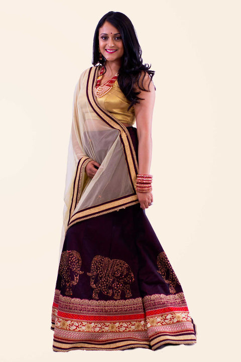 Exquisite eggplant skirt with gold elephant embroidery on multicolor border. Paired with gold blouse and color contrasting net dupatta for draping.